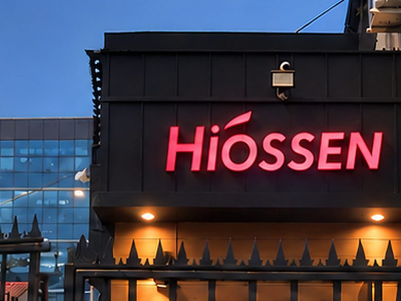 Muro del costado del acceso con el logotipo Hiossen iluminado en rojo sobre el revestimiento oscuro